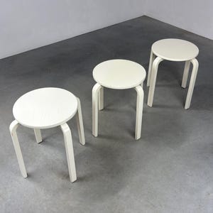 Könnte beinhalten: Drei weiße Holzstühle mit runden Sitzflächen und gebogenen Beinen. Sie stehen in einem Raum mit grauem Boden und Wänden. Das Design ist minimalistisch. Die Sitzflächen haben einen Durchmesser von etwa 30 cm.