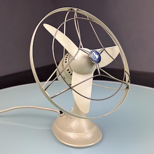 Peut inclure: Un ventilateur électrique vintage blanc cassé avec trois pales, une cage de protection et une base ronde. Le ventilateur a un emblème bleu et blanc à l'avant et un cordon d'alimentation. Le ventilateur est sur une surface bleu clair.