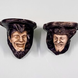 Puede incluir: Dos soportes decorativos para estantes de pared hechos de yeso. Cada soporte presenta una cara esculpida con una expresión caprichosa. Una cara sonríe con una amplia sonrisa, mientras que la otra tiene una expresión más seria. Los soportes están pintados en un color marrón oscuro con acentos blancos.