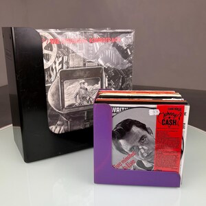 May include: A black vinyl record storage case and a purple vinyl record holder. The purple holder displays a Johnny Cash album cover. The black case contains a soundtrack album. The albums are on a white surface.