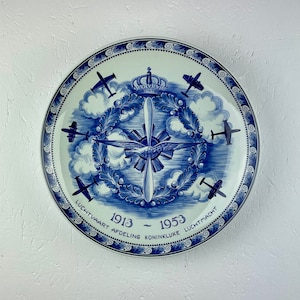 Puede incluir: Un plato de cerámica azul y blanco con un diseño de aviones y una corona. El texto en el plato dice "Luchtvaart Afdeling Koninklijke Luchtmacht 1913 - 1953".