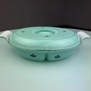 Może przedstawiać: W stylu vintage, miętowo-zielone naczynie do zapiekania z emalii z pokrywką i dwoma uchwytami. Naczynie posiada kwiatowy wzór i białe paski na uchwytach. To retro naczynie kuchenne jest idealne do serwowania i przechowywania żywności.