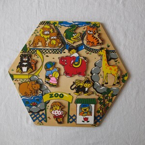 Può includere: Un puzzle di legno a forma di esagono con tema zoo. I pezzi del puzzle includono una giraffa, un ippopotamo, una tigre, un leone, un elefante, un orso nero, una scimmia e diverse persone. Il puzzle è dipinto con colori vivaci e ha un cartello "ZOO".
