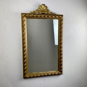 Puede incluir: Espejo rectangular con marco dorado y parte superior decorativa en forma de concha. El marco tiene una textura de cuerda y un borde detallado. El espejo está colgado en una pared blanca.