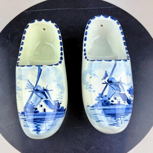 Könnte beinhalten: Ein Paar cremefarbene Keramik-Holzschuhe mit einer handgemalten blauen Windmühlenszene auf der Vorderseite. Die Ränder der Öffnungen sind mit einem wiederkehrenden blauen Punktmuster verziert. Jeder Schuh hat ein kleines Loch am Fersenbereich.