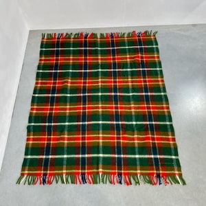 May include: A rectangular plaid blanket in green, red, and navy blue. The blanket has a fringed edge with green, red, and blue threads. The plaid pattern is a classic design, suitable for various uses. The blanket is likely made of wool or a similar material.