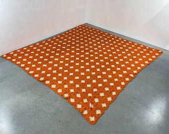 May include: A large, orange and white crocheted blanket with a square pattern. The blanket is laid out on a gray concrete floor.