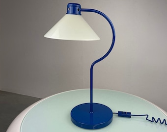 May include: A blue desk lamp with a white shade. The lamp has a curved arm and a round base. The lamp is on a white table.