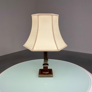 Op de afbeelding: Een witte tafellamp met een zeshoekige lampenkap staat op een ronde, lichtblauwe tafel. De lamp heeft een gouden voet en een bruine accent.