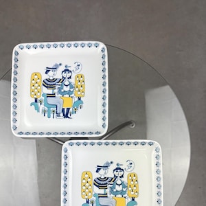 Op de afbeelding: Twee vierkante keramische borden met een blauwe en witte rand. In het midden van elk bord staat een gestileerde illustratie van een stel dat op een bankje zit met een vogel en een halve maan op de achtergrond. Het stel draagt blauwe en gele kleding.