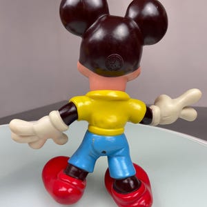 May include: A vintage plastic Mickey Mouse figurine with brown ears, a yellow shirt, blue pants, and red shoes. The figurine has a brown face and white gloves.