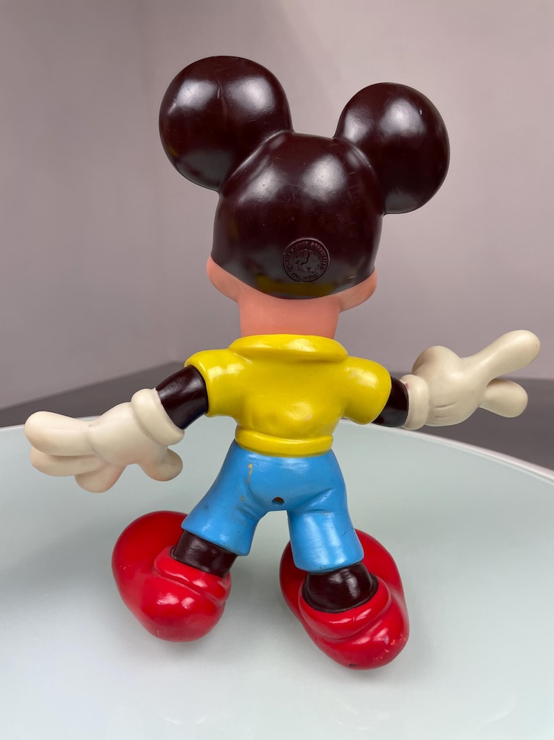 May include: A vintage plastic Mickey Mouse figurine with brown ears, a yellow shirt, blue pants, and red shoes. The figurine has a brown face and white gloves.