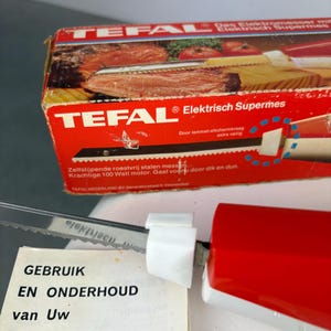 May include: A red and white TEFAL electric carving knife set. The box features the TEFAL logo and the words "Elektrisch Supermes". The knife has a serrated stainless steel blade and a red handle. The box also includes the text "GEBRUIK EN ONDERHOUD".