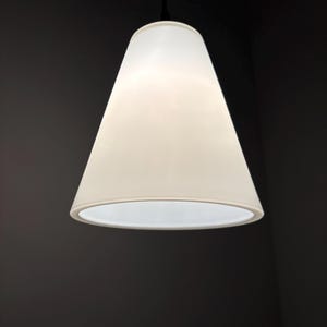 Op de afbeelding: Een witte, kegelvormige hanglamp hangt aan een zwart snoer tegen een donkergrijze achtergrond. Het licht is aan en verlicht de binnenkant van de kap. De armatuur heeft een eenvoudig, modern ontwerp.