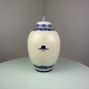 Op de afbeelding: Een witte keramische pot met een blauw-wit bloemenpatroon en een blauwe hoed met het nummer 1616 erop. De pot heeft een deksel met een klein blauw-wit bloemenpatroon.