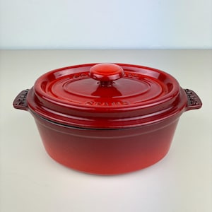 May include: Red oval cast iron Dutch oven with a lid and a knob. The oven has a gradient color from dark red to light red.