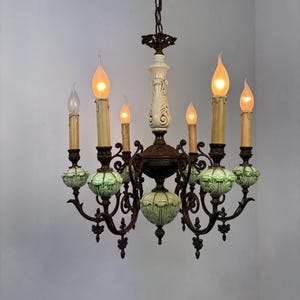 May include: An antique chandelier with six candle-style lights. The fixture features a dark bronze frame, cream-colored accents, and green ceramic globes. The bulbs are lit, casting a warm glow. The chandelier hangs from a chain.
