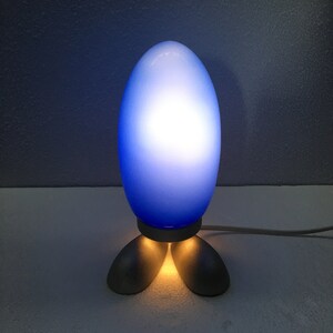 May include: A blue glass lamp shaped like an egg with a white light shining through. The lamp has three silver feet.