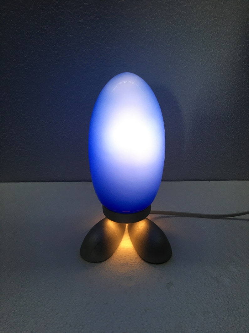 May include: A blue glass lamp shaped like an egg with a white light shining through. The lamp has three silver feet.