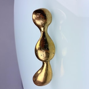 May include: A vertical cabinet handle made of polished brass with a smooth, organic, and irregular shape consisting of three connected rounded segments. The handle is mounted onto a smooth, light-colored, opaque surface with a glossy finish.