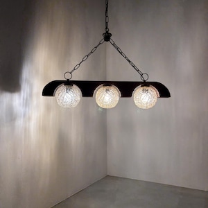 Op de afbeelding: Een lineaire hanglamp met een donkerbruine houten balk die aan zwarte metalen kettingen hangt. Aan de onderkant zijn drie bolvormige glazen kappen met een getextureerde, gebarsten afwerking bevestigd, elk met een verlichte gloeilamp.