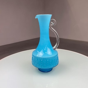Op de afbeelding: Een vintage turquoise glazen kan met een helder, gedraaid handvat. De kan heeft een getextureerd lichaam en een decoratieve basis met een reliëfpatroon. De kan wordt weergegeven op een wit oppervlak.