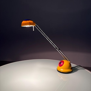 Op de afbeelding: Een gele en blauwe bureaulamp met een rode knop op de basis. De lamp heeft een flexibele arm en een koepelvormige kap.