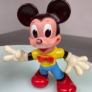 May include: A vintage plastic Mickey Mouse figurine with large black ears, a yellow shirt, a red bow tie, blue pants, and red shoes.