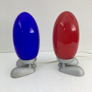 May include: Two table lamps with a unique design. The lamps have a rounded, egg-shaped shade and three silver legs. One lamp has a blue shade and the other has a red shade.