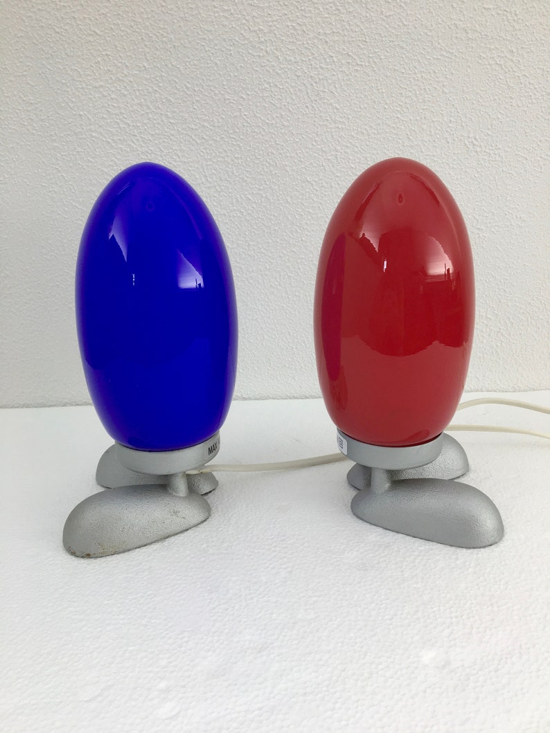 May include: Two table lamps with a unique design. The lamps have a rounded, egg-shaped shade and three silver legs. One lamp has a blue shade and the other has a red shade.