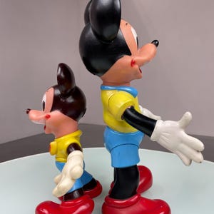 May include: Two plastic Mickey Mouse figurines, one larger than the other. Both are wearing yellow shirts, blue pants, and red shoes. The larger Mickey Mouse is smiling and has his arms outstretched.