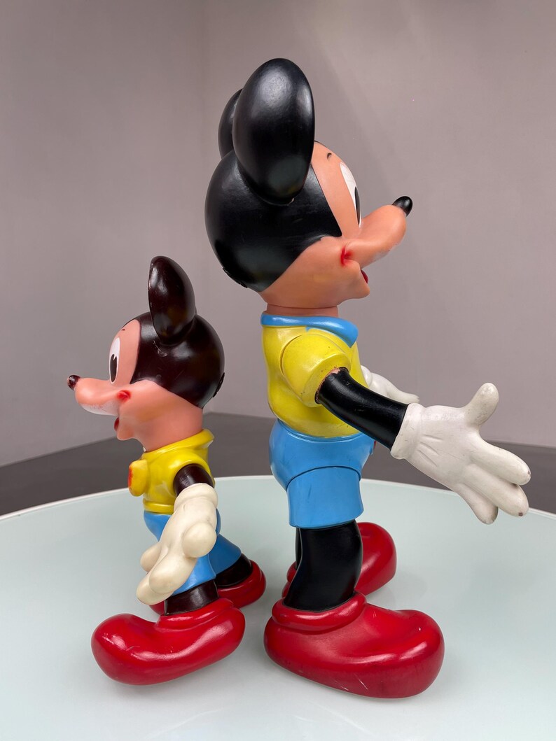 May include: Two plastic Mickey Mouse figurines, one larger than the other. Both are wearing yellow shirts, blue pants, and red shoes. The larger Mickey Mouse is smiling and has his arms outstretched.