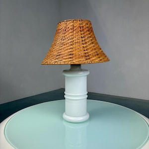 Op de afbeelding: Een vintage tafellamp met een geweven rotan lampenkap. De lamp heeft een witte, cilindrische voet met decoratieve ringen. De lamp staat op een ronde tafel met een lichtblauw glazen blad. De lamp is een decoratieve verlichtingsarmatuur.