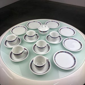 Op de afbeelding: Een wit porseleinen theeservies met een decoratieve blauwe, paarse en roze rand. De set bevat kopjes, schoteltjes, kleine bordjes en een suikerpot met een deksel. De items zijn gerangschikt op een ronde, lichtgroene tafel.