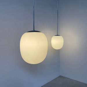 Könnte beinhalten: Zwei weiße Globus-Pendelleuchten, die an der Decke hängen, vor einer hellblauen Wand. Die größere Kugel befindet sich im Vordergrund und die kleinere Kugel im Hintergrund.