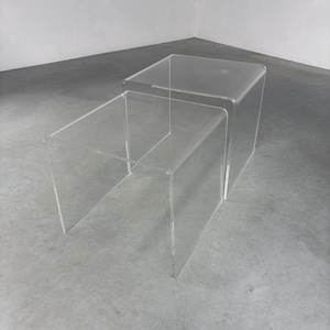 May include: Two clear acrylic nesting tables. The larger table is on top of the smaller table. Both tables have a square top and four legs.