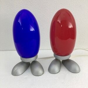 May include: Two table lamps with a unique design. The lamps have a rounded, egg-shaped base with a silver finish and three legs. The top of the lamps are a bright blue and a red.