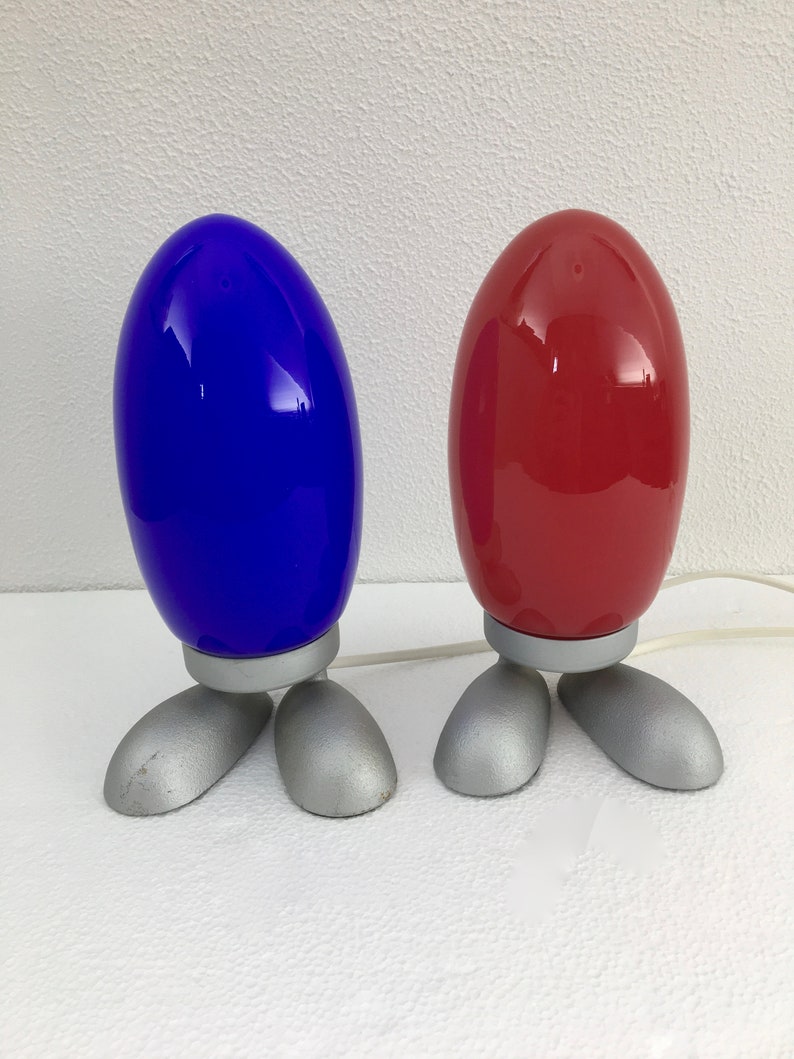 May include: Two table lamps with a unique design. The lamps have a rounded, egg-shaped base with a silver finish and three legs. The top of the lamps are a bright blue and a red.