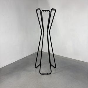 May include: A black metal coat rack with a minimalist design. The rack has a tall, slender frame with a series of curved hooks at the top for hanging coats, hats, or other items. The base has a curved design. The rack is approximately 60 inches tall.