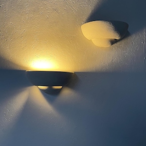 May include: Two white wall sconces with a textured finish. The top sconce is off and the bottom sconce is on, casting a warm yellow light on the wall.