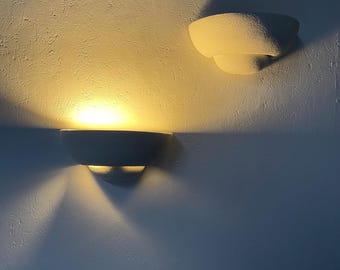 May include: Two white wall sconces with a textured finish. The top sconce is off and the bottom sconce is on, casting a warm yellow light on the wall.