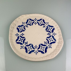 Peut inclure: Une assiette ronde en céramique blanc cassé avec un motif floral bleu. L'assiette présente un bord festonné et un motif central de fleurs bleues stylisées. Posée sur un fond bleu clair.
