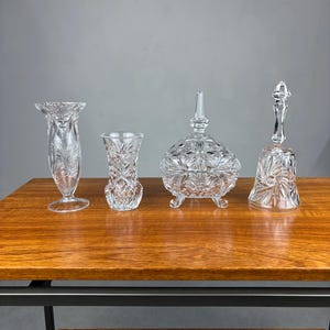 Op de afbeelding: Vier heldere kristallen decoratieve items op een houten tafel. De items omvatten twee vazen, een afgedekte bonbonschaal en een bel. De kristallen stukken hebben ingewikkelde snijpatronen.