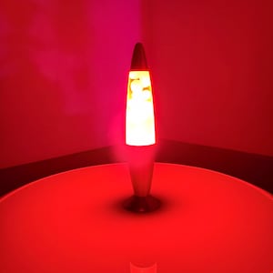 Op de afbeelding: Een lavalamp met een conische bovenkant en een ronde voet. De glazen houder bevat een heldere witte vloeistof met zwevende witte wasvormen. De lamp geeft een sterk rood schijnsel af en verlicht de omgeving met een levendig rood licht.