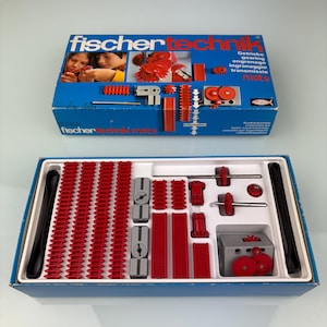 Peut inclure: Un jeu de construction Fischertechnik mot 2 vintage. La boîte bleue présente le nom de la marque et des images d'enfants. À l'intérieur, des composants en plastique rouge et gris, dont des engrenages, des chenilles et des connecteurs, sont soigneusement rangés.