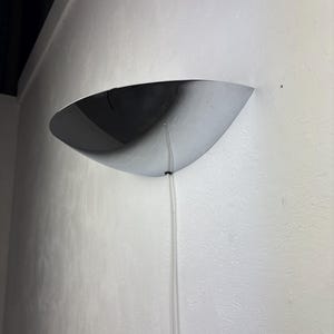 May include: A contemporary wall sconce with a chrome finish, shaped like a half-moon. It's mounted on a textured white wall, with a clear cable hanging down. The sconce has a reflective surface.
