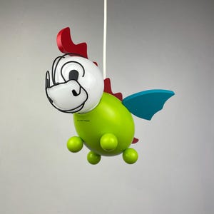 Könnte beinhalten: Eine grüne und rote drachenförmige Pendelleuchte mit weißem Gesicht und blauen Flügeln. Die Leuchte hängt an einem weißen Kabel.