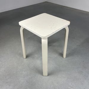 Puede incluir: Un taburete de madera blanco con cuatro patas y una parte superior cuadrada. El taburete tiene un acabado desgastado, lo que le da un aspecto vintage.