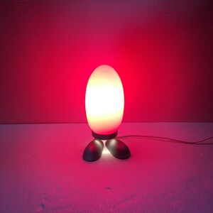 May include: A white egg-shaped lamp with a red glow, standing on two black feet. The lamp is on a white surface.