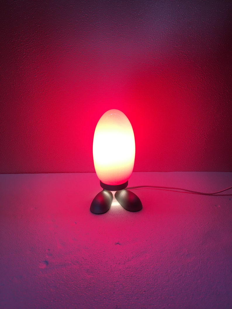 May include: A white egg-shaped lamp with a red glow, standing on two black feet. The lamp is on a white surface.